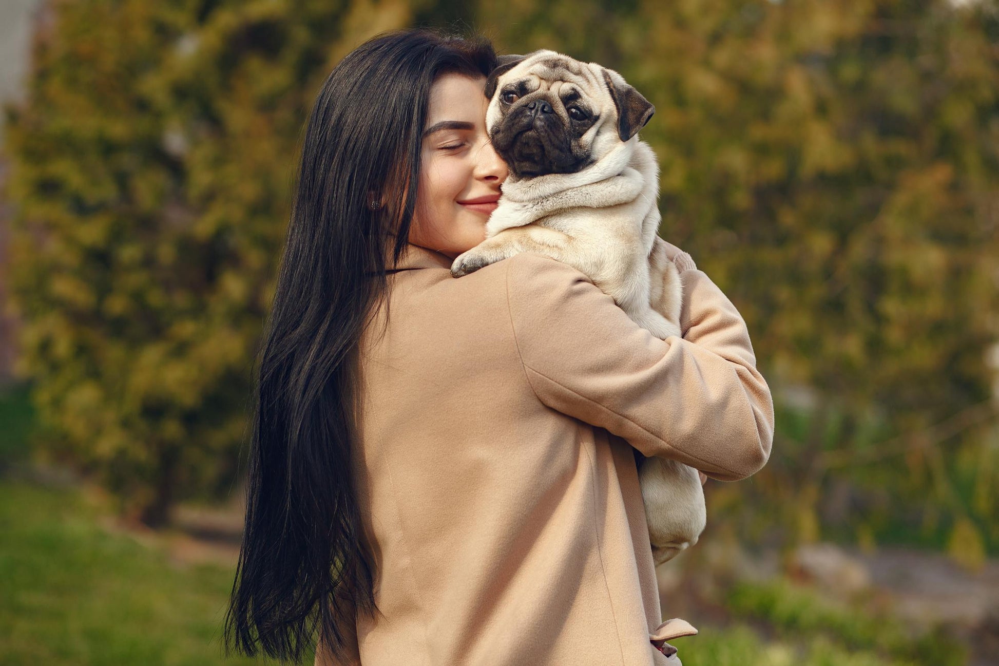 Woman holding and snuggling a pug in a green meadow with trees. 